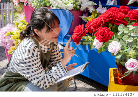 A happy female flower shop employee is working in the garden of her shop, checking flower stocks. 111124107