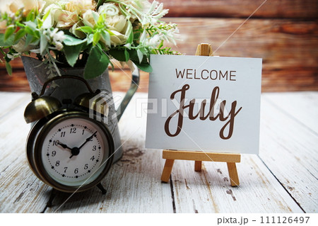 Welcome July text message written on paper card with wooden easel and alarm clock with flower in metal vase decoration Welcome July text message written on paper card with wooden easel and alarm clock with flower in metal vase decoration 111126497