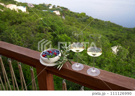 White wine in glasses against a background of blue sky and sea and a plate with raspberries and blueberries next to it . High angle. Copy Space. 111126990