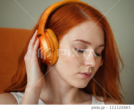 Close-up portrait of a young red-haired woman wearing orange headphones. 111128865
