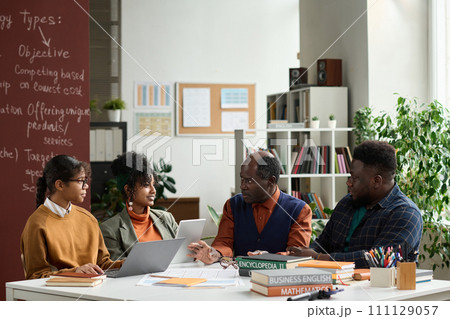 Portrait of senior Black professor talking to group of students sitting at table together in college classroom copy space Portrait of senior Black professor talking to group of students sitting at table together in college classroom copy space 111129057