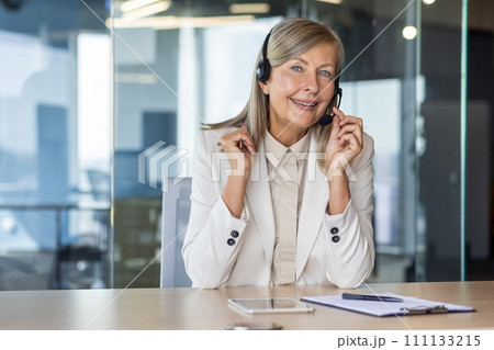 Happy elderly female in light formal clothes in wireless headset setting microphone by table in office space. Blue eyed woman working as customer support representative and providing service. 111133215