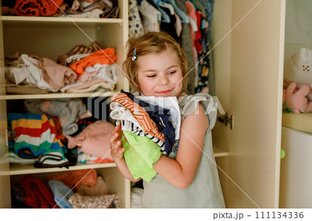 Little school girl standing by wardrobe with clothes. Preschool child putting shirts away and making decision for shirt to wear. Children get dressed in the morning for school. 111134336