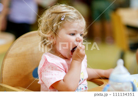 Adorable toddler girl eating healthy vegetables and unhealthy french fries potatoes. Cute happy baby child taking food from dish at daycare or nursery canteen. 111134351