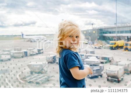 Cute little toddler girl at the airport, traveling. Happy healthy child waiting near window and watching airplanes. Family going on summer vacations by plane. 111134375