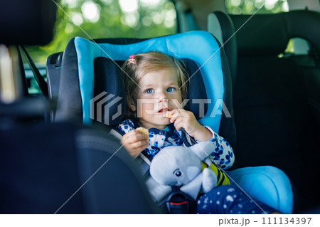 Adorable baby girl with blue eyes sitting in car safety seat. Toddler child going on family vacations and jorney. Smiling happy child during traffic jam, eating bisquit 111134397