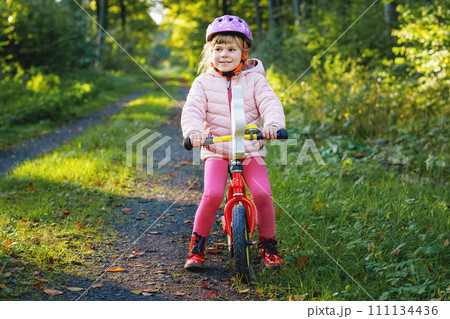 Child riding balance bike. Kids on bicycle in sunny forest. Little girl enjoying to ride glider bike on warm day. Preschooler learning to balance on run bicycle in safe helmet. Sport activity Child riding balance bike. Kids on bicycle in sunny forest. Little girl enjoying to ride glider bike on warm day. Preschooler learning to balance on run bicycle in safe helmet. Sport activity 111134436