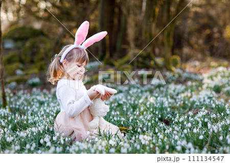 Adorable little girl with Easter bunny ears holding soft plush toy in spring forest on sunny day, outdoors. Cute happy child with lots of snowdrop flowers. Springtime, christian holiday concept. Adorable little girl with Easter bunny ears holding soft plush toy in spring forest on sunny day, outdoors. Cute happy child with lots of snowdrop flowers. Springtime, christian holiday concept. 111134547