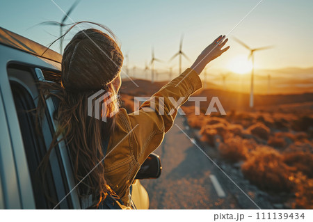A joyful woman looks at wind generators and the sunset from a car window. A joyful woman looks at wind generators and the sunset from a car window. 111139434