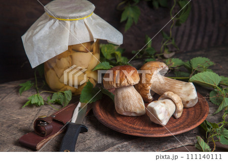 Clay plate with porcini mushroom commonly known as Boletus Edulis, glass jar with canned mushrooms and knife on vintage wooden background.. 111140111