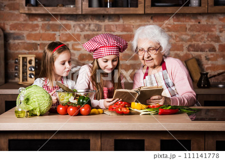 Granny with two granddaughters reading recipe from cooking book and cooking salad Granny with two granddaughters reading recipe from cooking book and cooking salad 111141778