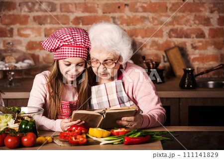 Little granddaughter reading recipe book with her granny sitting at kitchen Little granddaughter reading recipe book with her granny sitting at kitchen 111141829