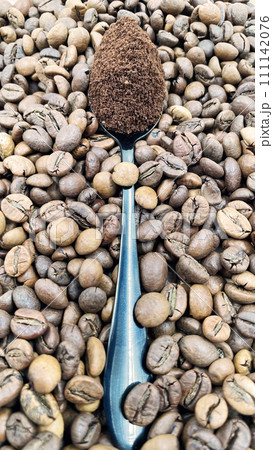 Against the background of roasted aromatic coffee beans lies a metal spoon filled with ground coffee. A drink made from roasted and ground beans from the coffee tree or coffee bush Against the background of roasted aromatic coffee beans lies a metal spoon filled with ground coffee. A drink made from roasted and ground beans from the coffee tree or coffee bush 111142076