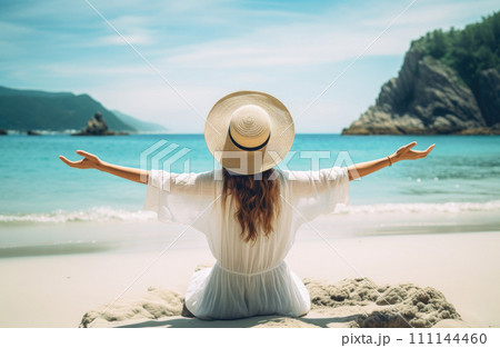 Young woman in white dress and hat sitting on the beach 111144460