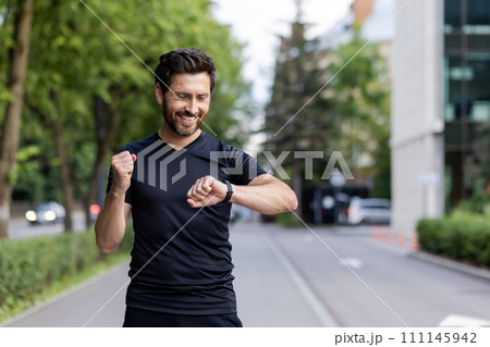 Happy young man doing sports and running, standing on city street, looking at wrist tracker and showing success gesture with hand. 111145942