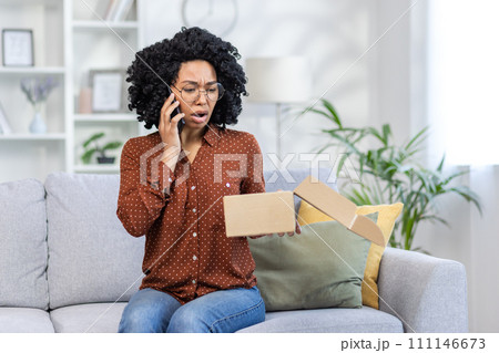 Angry African American young woman talking on the phone. He sits on the sofa at home and anxiously looks at the open box of the package he received in the mail. 111146673