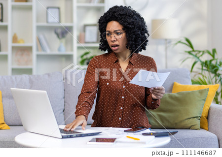 Worried young African American woman sitting on sofa at home and working on laptop, holding bills and documents, checking data online. 111146687