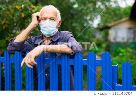 Senior man in mask leaning on fence and looking in camera 111146704