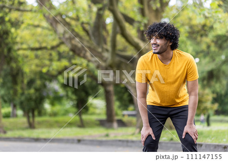 Tired and satisfied with himself and the result, an Indian young man rests after a run, smiling, bent over and looking to the side. Tired and satisfied with himself and the result, an Indian young man rests after a run, smiling, bent over and looking to the side. 111147155