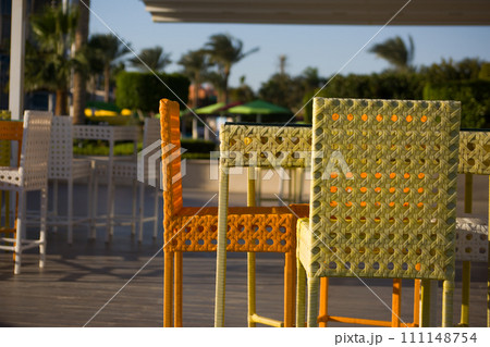 Back of a wicker chair at a table in a cafe by the sea. Beach cafe against the backdrop of a park with plants and flowers Back of a wicker chair at a table in a cafe by the sea. Beach cafe against the backdrop of a park with plants and flowers 111148754