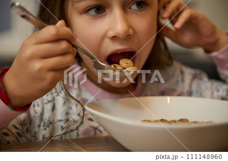 Close-up face portrait of hungry child girl holding full table spoon of sweet delicious corn oat flakes with milk honey 111148960