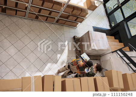 Top view of workers checking customers orders on laptop before packing and shipping, working in storage room. Diverse team weraring protective overall overseeing online production of merchandise. Top view of workers checking customers orders on laptop before packing and shipping, working in storage room. Diverse team weraring protective overall overseeing online production of merchandise. 111150248