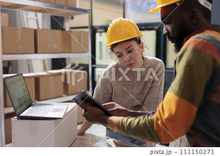 Diverse coworkers checking online orders on tablet computer, analyzing products for inventory report in warehouse. Stockroom team working at customers packages preparing transportation 111150271