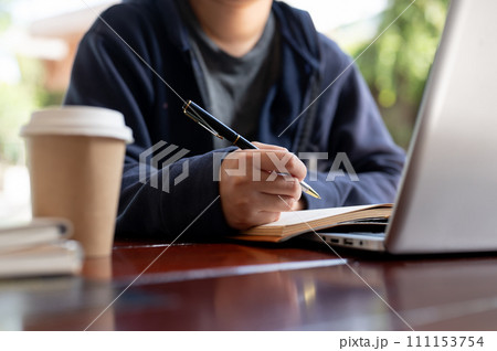 A woman is jotting down something in a notebook while working on her laptop at a table in a cafe. 111153754