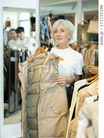 Senior woman chooses vest on hangers and closely examines its details, feels texture of fabric. 111155296