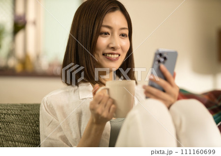 ソファーでくつろいでコーヒーを飲みながらスマホを見るミドル女性 111160699