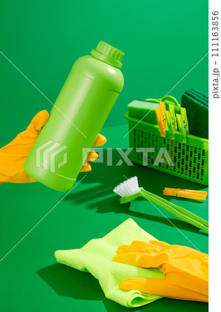 Cleaner with yellow rubber gloves holding unlabeled detergent bottle on green background. In the back is a plastic basket with cleaning supplies. House cleaning concept, photo for advertising Cleaner with yellow rubber gloves holding unlabeled detergent bottle on green background. In the back is a plastic basket with cleaning supplies. House cleaning concept, photo for advertising 111163856