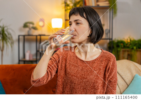 Smiling Caucasian woman sitting at home leading healthy lifestyle, drinking a glass of pure water 111165400