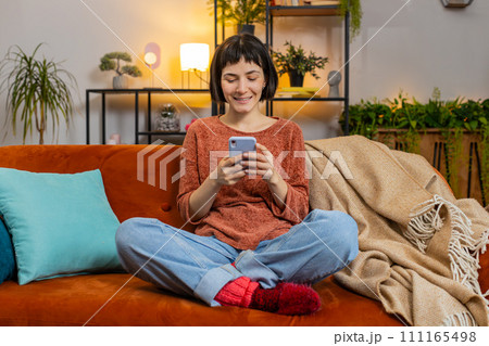 Happy woman thinking texting messaging on smartphone watching online movie sitting on home sofa Happy woman thinking texting messaging on smartphone watching online movie sitting on home sofa 111165498