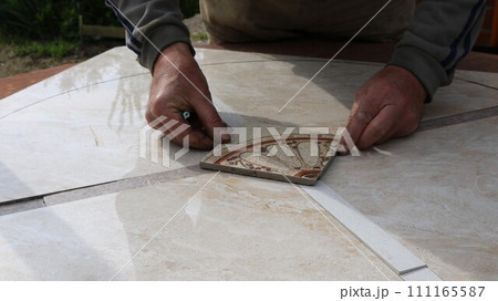 The tiler's hands place a square piece of material with a floral pattern on the smooth surface of a large light-colored tile, assembling a mosaic when tiling a table surface The tiler's hands place a square piece of material with a floral pattern on the smooth surface of a large light-colored tile, assembling a mosaic when tiling a table surface 111165587