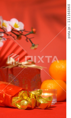 Chinese Lunar New Year red background. ancient Chinese ingot gold bar in silk bag, red gift box with golden bow, paper fan, oranges, plum blossom branch, candle and foliage shadow sway. vertical Chinese Lunar New Year red background. ancient Chinese ingot gold bar in silk bag, red gift box with golden bow, paper fan, oranges, plum blossom branch, candle and foliage shadow sway. vertical 111168868