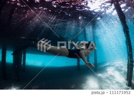 Freediver lady swimming underwater under the pier in blue ocean with sun rays. 111171952