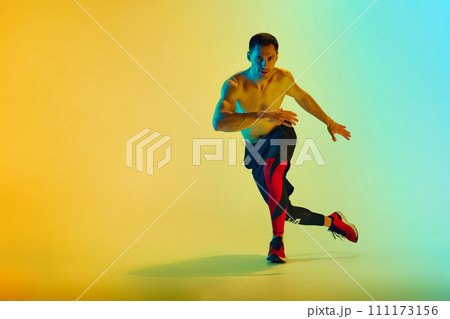 Shirtless male athlete in mid-action with focused expression training shirtless against gradient blue yellow background in neon light 111173156