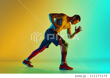 Athletic young man, with muscular, relief shirtless body training with resistance band against gradient blue yellow background in neon light Athletic young man, with muscular, relief shirtless body training with resistance band against gradient blue yellow background in neon light 111173174