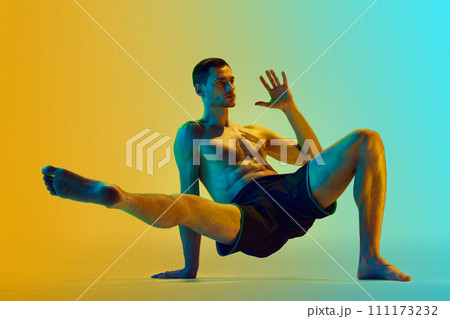 Intensive full-body training. Muscular athletic young shirtless man, with fit body doing workout exercises against gradient blue yellow background in neon light 111173232