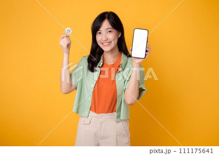 future finance with a cheerful young Asian woman in her 30s, donning orange shirt and green jumper, showcasing smartphone screen display and crypto currency coin on yellow background. future finance with a cheerful young Asian woman in her 30s, donning orange shirt and green jumper, showcasing smartphone screen display and crypto currency coin on yellow background. 111175641
