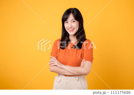 Embody confidence and poise with a young Asian woman in her 30s, donning an orange shirt, showcasing crossed arm sign gesture on yellow background. 111175686
