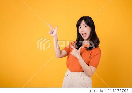 Spread happiness with a young Asian woman in her 30s, donning an orange shirt, pointing to free copy space on yellow background. recommending product concept. 111175702