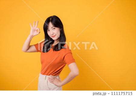 Embody joy with a young Asian woman in her 30s, donning an orange shirt, giving a okay hand gesture on yellow background. product recommending concept. Embody joy with a young Asian woman in her 30s, donning an orange shirt, giving a okay hand gesture on yellow background. product recommending concept. 111175715