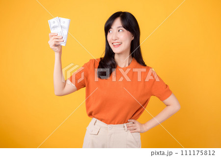 Lively young Asian woman in her 30s, holding cash money dollars, standing on vibrant yellow backdrop. Lively young Asian woman in her 30s, holding cash money dollars, standing on vibrant yellow backdrop. 111175812