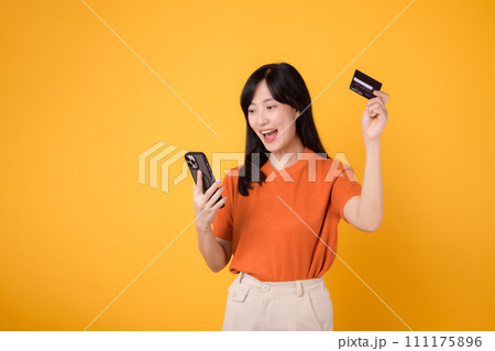 Cheerful Asian woman 30s in orange shirt, using smartphone and holding credit card on yellow background. Convenient online shopping. 111175896