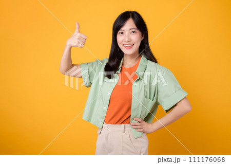 positive feedback of approval with young Asian woman 30s, dressed gracefully in orange shirt and green jumper. Her expressive thumbs up gesture, captured on yellow background, concept of acceptance. 111176068