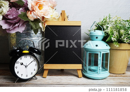Easel mockup with blue landtern, alarm clock and flowers bouquet decoration on wooden background 111181858