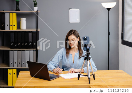 Videoconference And Training. Class of diverse students learning remotely online using web app. Female teacher providing lesson with laptop, recording video on cellphone on tripod Videoconference And Training. Class of diverse students learning remotely online using web app. Female teacher providing lesson with laptop, recording video on cellphone on tripod 111184216