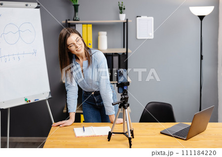 Positive blogger woman speaking at smartphone webcam on tripod, recording post for vlog. Online teacher, coach working, giving webinar at home. Presenter taking video on cellphone Positive blogger woman speaking at smartphone webcam on tripod, recording post for vlog. Online teacher, coach working, giving webinar at home. Presenter taking video on cellphone 111184220