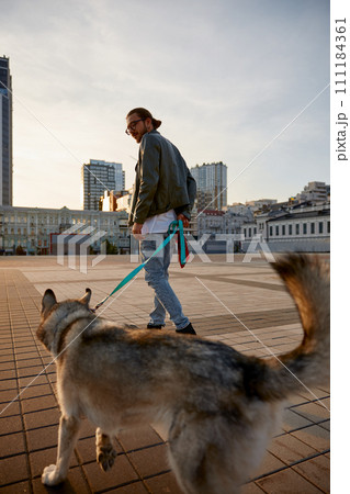 Hipster guy skateboarding and walking dog moving together through city square Hipster guy skateboarding and walking dog moving together through city square 111184361